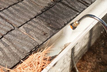 "A house rain gutter is being cleaned with a specialized curved pressure washer nozzle. The bottom left portion of the gutter is filled with pine needles and debris, the right gutter is clean."