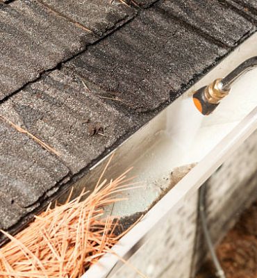 "A house rain gutter is being cleaned with a specialized curved pressure washer nozzle. The bottom left portion of the gutter is filled with pine needles and debris, the right gutter is clean."